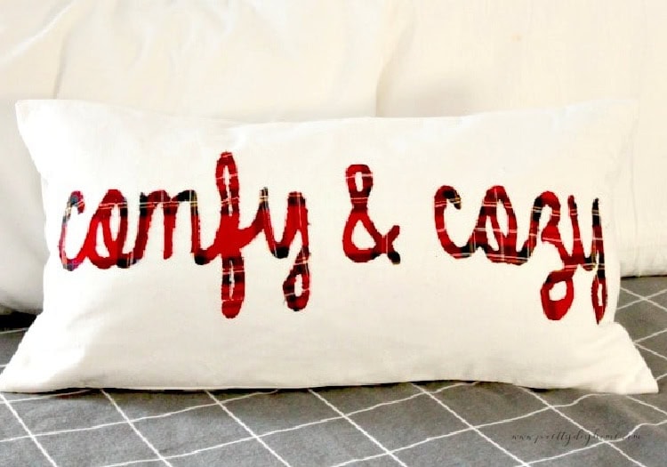 Christmas pillows DIY with red plaid lettering that says comfy and cozy.  The cushion cover is bright white, oblong and sitting on a grey flannel bedspread.