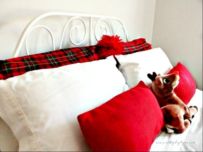 The upper portion of a bed decorated for Christmas with two red tartan DIY fuzzy  large cushion covers with big red pom poms.  Then two white pillow cases and then two small oblong red DIY envelope cushions.  