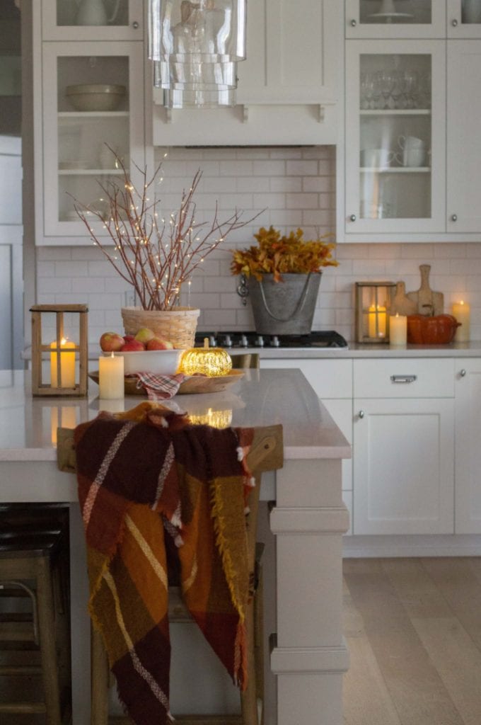 A cozy kitchen decorated for fall with lights.. There are branches with lights, and lanterns with lights along white countertops and cabinets.