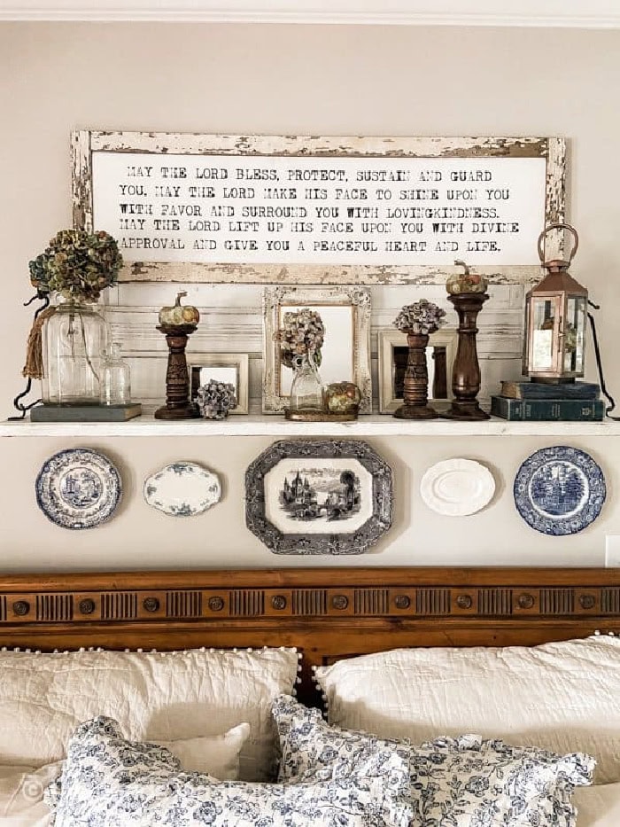 A antique mantel being repurposed over a headboard.  The mantel is white and chippy and theur are several different vintage finds on top.  A lanter, books, vase, old frame and dried floral arrangments.  Its very vintage and feminine.