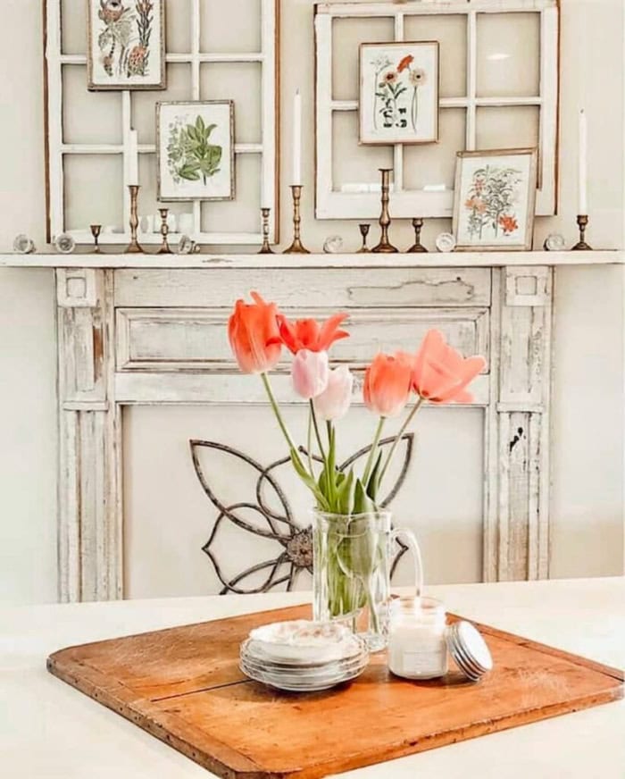 A vintage faux fireplace with old frames on the mantel, its sitting in a dining room with pretty flowers on the table.