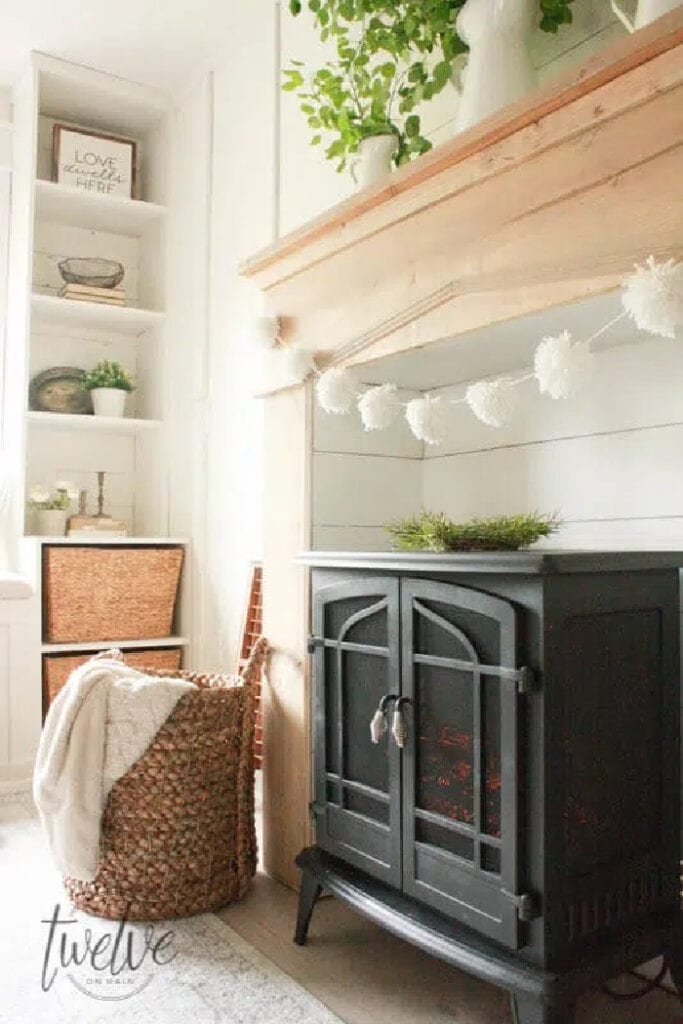 A handmade solid wood fireplace suerouns in light wood tones, it has white pom pom garland and a electric stove where the firebox would be