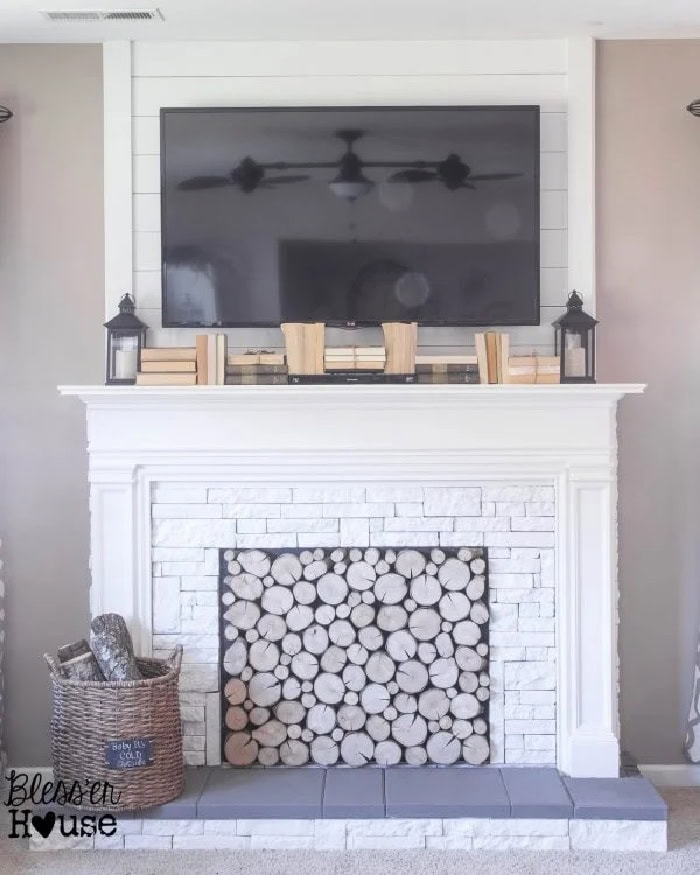 A large faux fireplace in bright white the upper part is shiplap with a tv, the lower part has a white mantel, white brick and the firebox is filled with cross cut log slices.