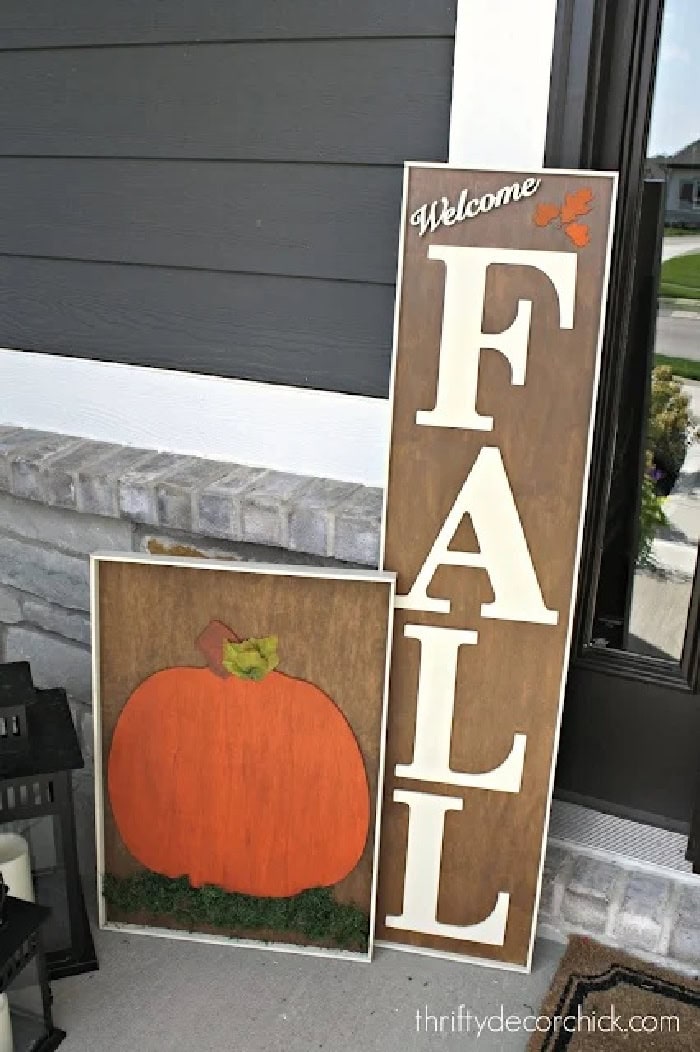 Two large fall DIY wood signs for the fall front porch. Both have white frames, and stained brown backgrounds. The short one had an orange pumpkin on the front the second tall one has the word FALL written on it.