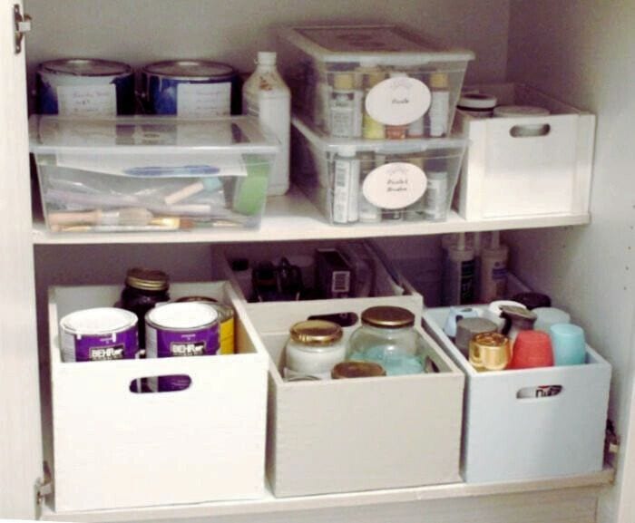 A cupboard filled with various pajnt supplies. In seeral different wood bins.  There are large paint cans, brushes, spray paints, small paints in clear jars and everything is labelled.