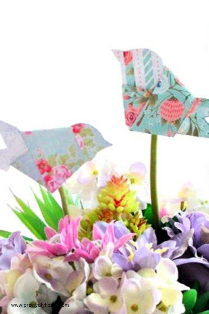 A close up of two pink and blue origami birds sitting on top of a Spring flower arrangement.