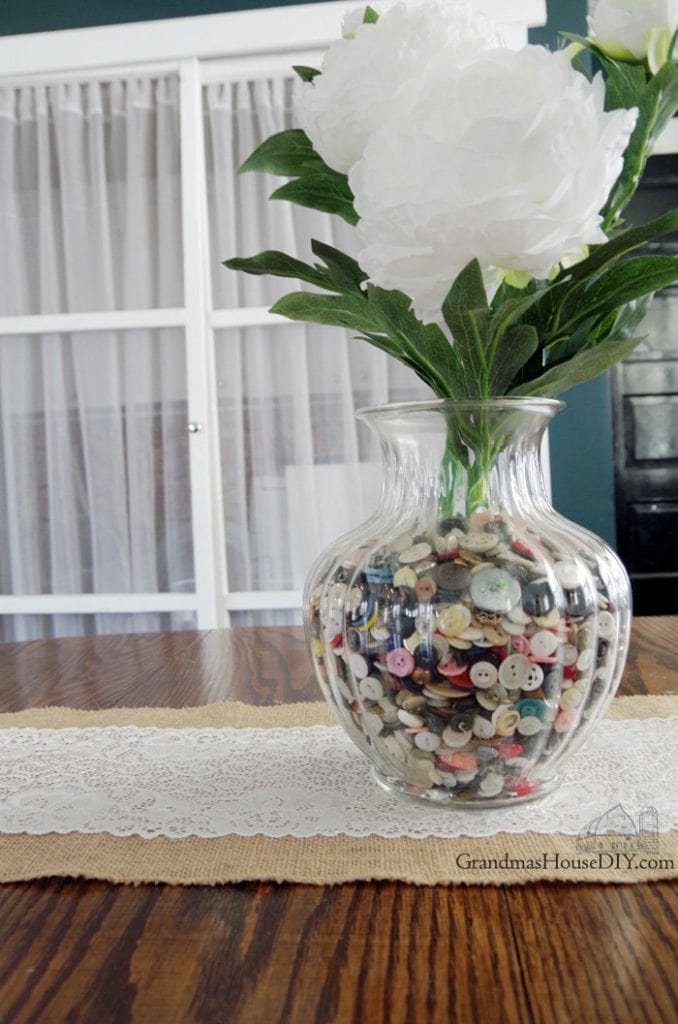 A large centerpiece of fresh peonies, in a clear glass vase filled with vintage buttons.