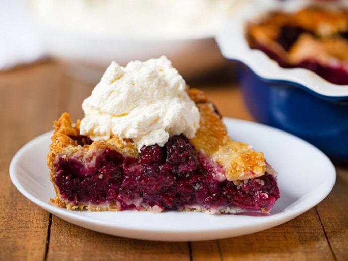 A slice of blackberry pie covered with a large dollop of whipped cream. The pie is served on a white plate and you can see the corner of the whole pie in the background.