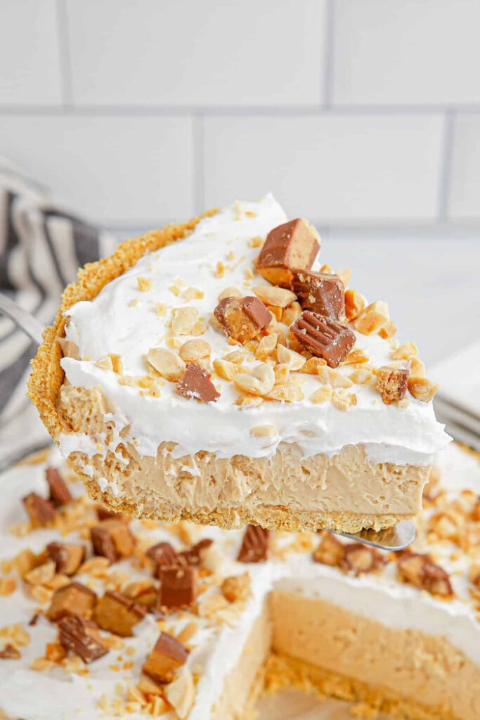 A slice of peanut butter pie showing a peanut butter cream cheese filling, a layer of whipped cream and topped with peanut chunks and chopped up reeces peanut butter cups.