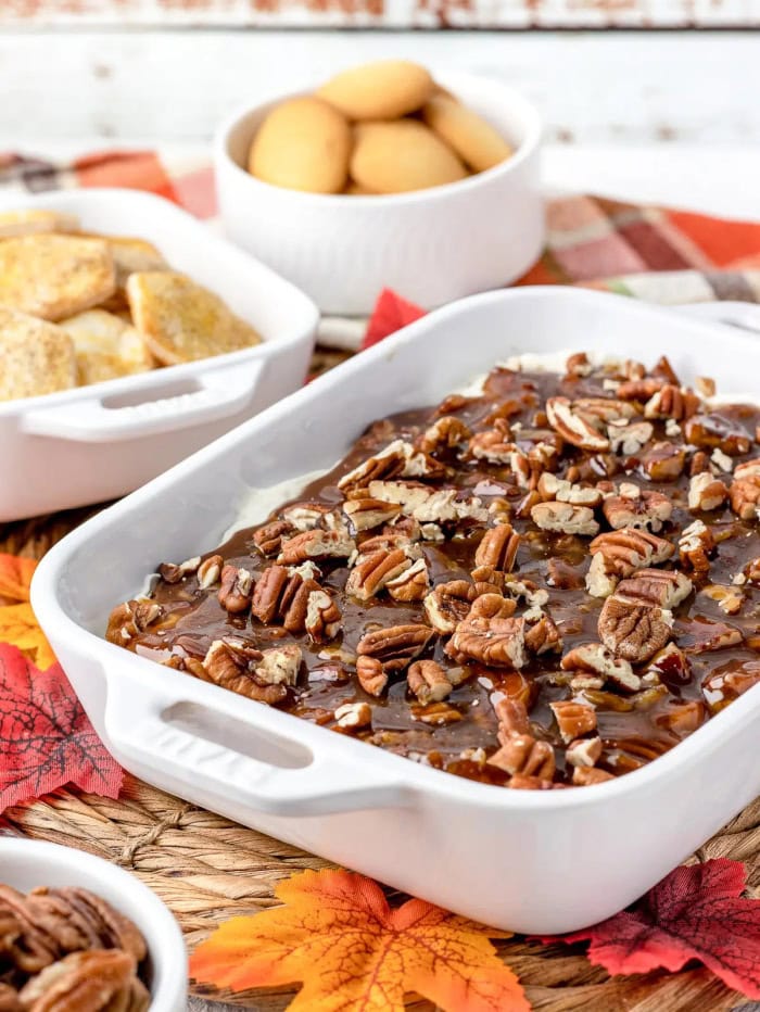 A small rectangular white baking dish filled with pecan pie dip with lots of pecans crushed on top. You can see two different bowls of plain cookies in the background for dipping.