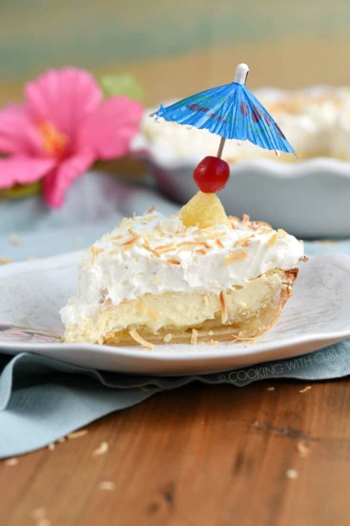 A pina colada flavored no bake pie recipe with a layer of custard filling whipped cream coconut and decorated with a pineapple marashino cherry on a blue drink umbrella.