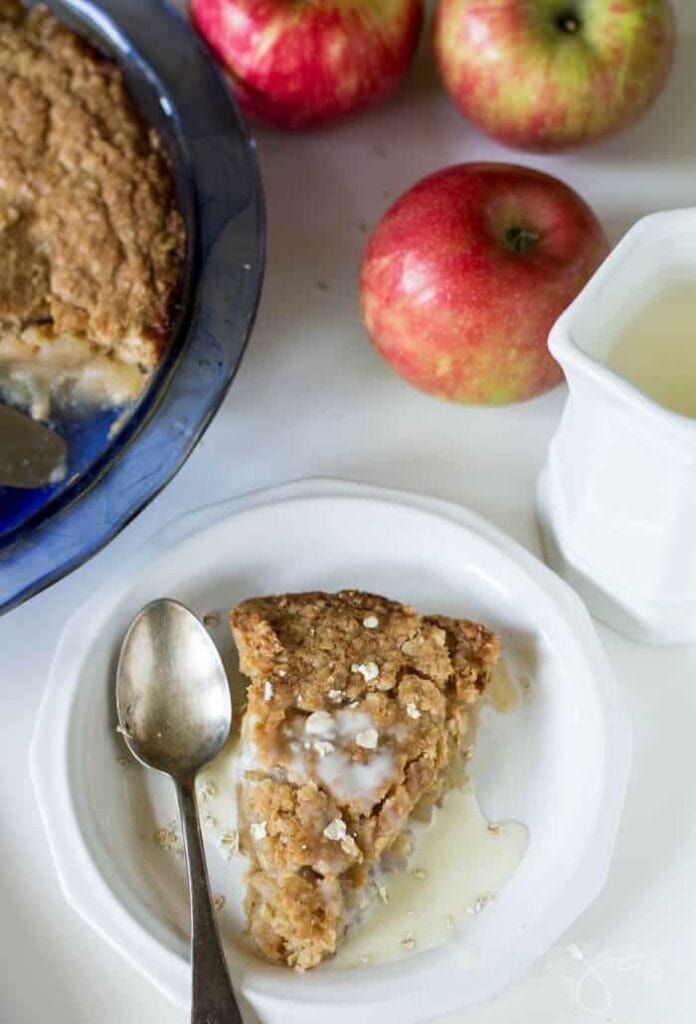 A pie without a bottom crust, filled with apples and covered with a oatmeal crumble. The pie is being served as a slice, and you can see a whole pie as well as red apples in the background.