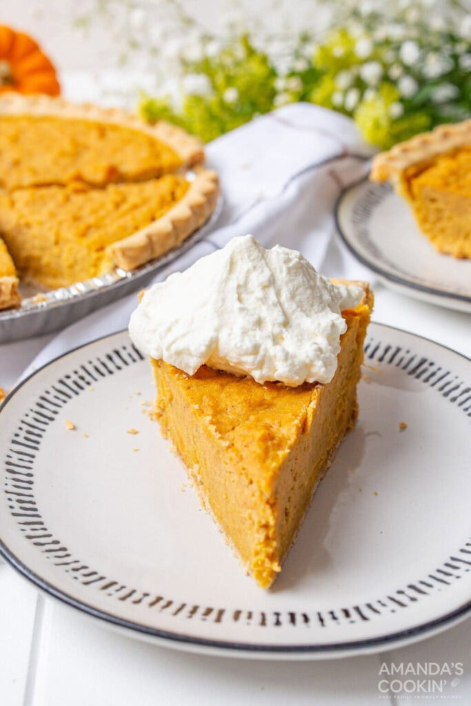 A thick solid slice of sweet potato pie with a dollop of whipped cream on top. You can see the whole pie in the background.