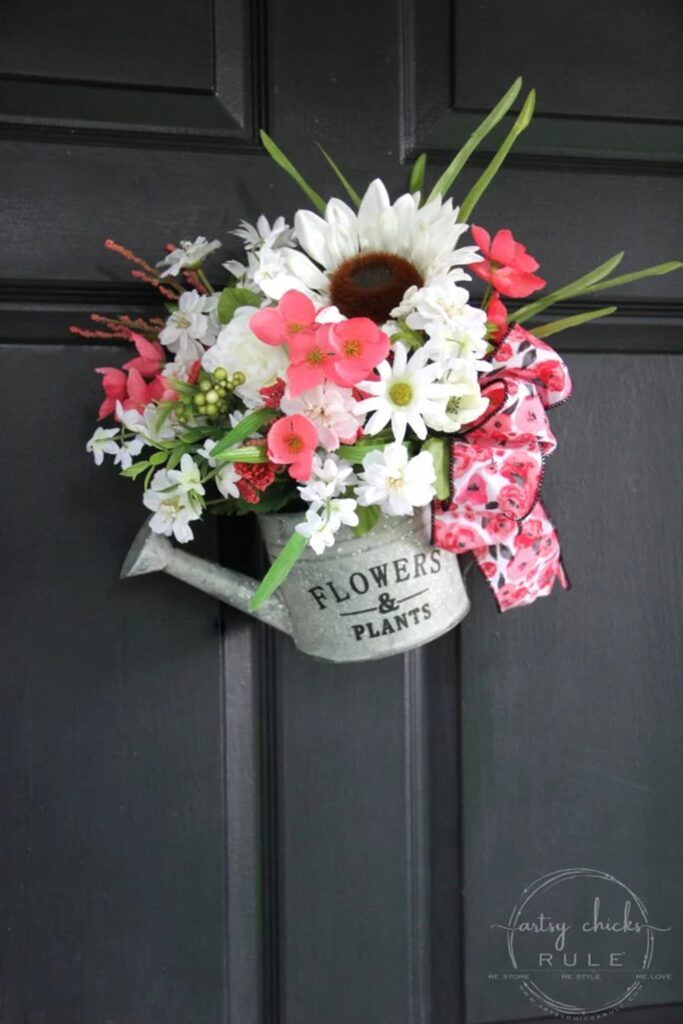 Repurposed watering can filled with flowers hanging on a black door for spring decor.