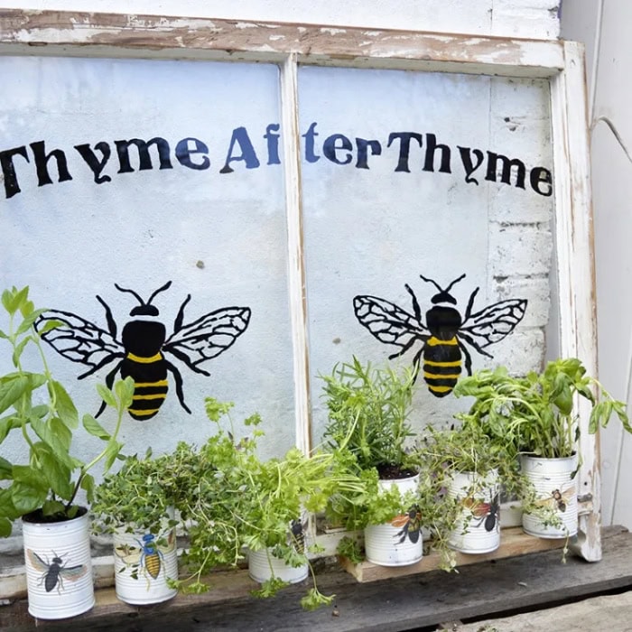 A repurposed window herb planter made with tin cans attached to an old window frame with a bee design.