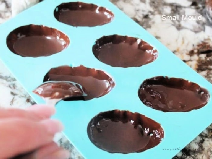 A small blue mold for chocolate Easter eggs, The mold is being filled with chocolate using a spoon, the interior is not filled.