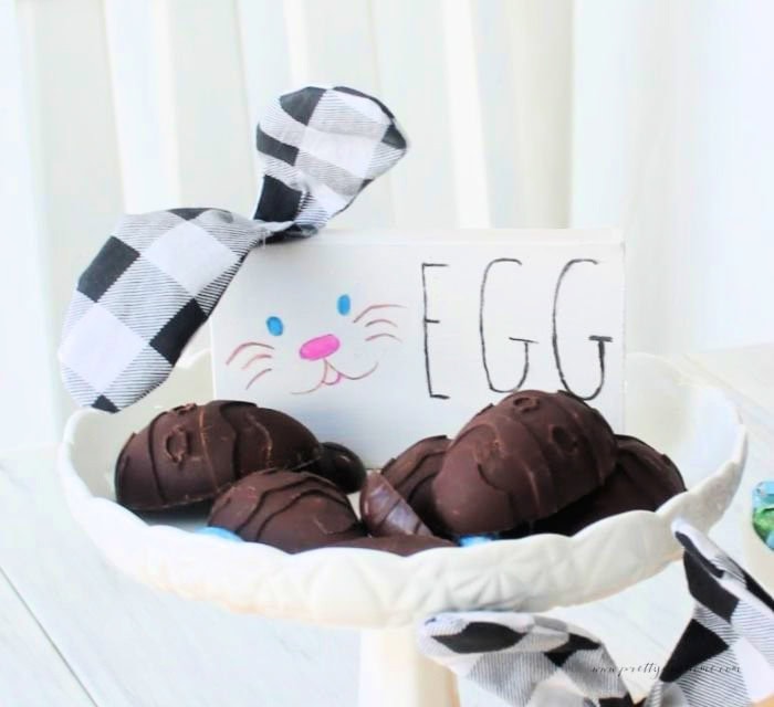 A white pedestal tray fulls of dark chocolate eater eggs.  There is a bunny sign with it that says Eggs.