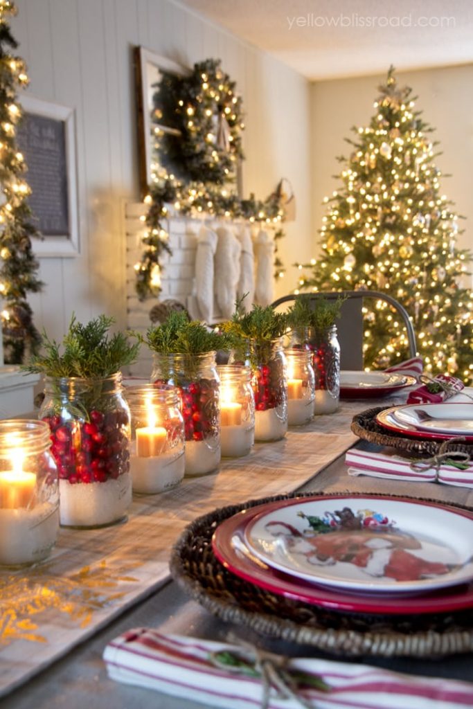 Table set with traditional red and green for Christmas with cranberries, sugar in jars, greenery and a tree in the background.