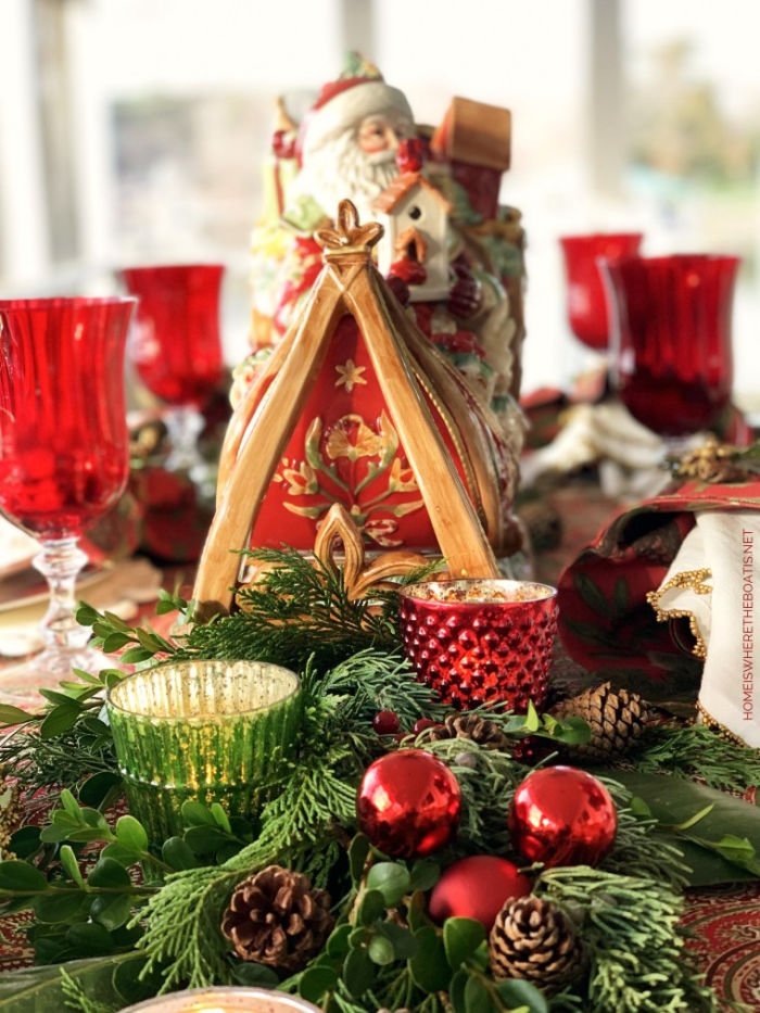 Traditional Christmas table setting with red balls, Santa centerpiece, candles, greenery, plaid tablecloth and red wine glasses.