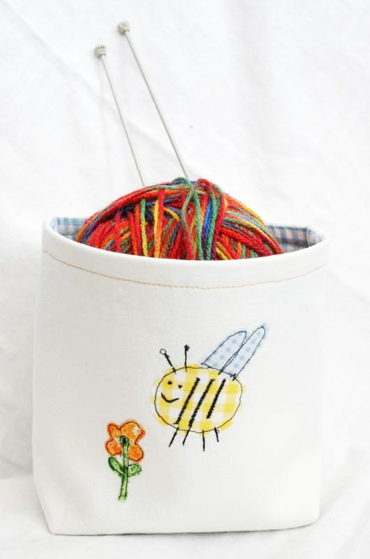 A sewn DIY fabric basket in cream with a fun bumblebee applique on the front. The DIY fabric bin has a ball of yarn inside with two knitting needles.