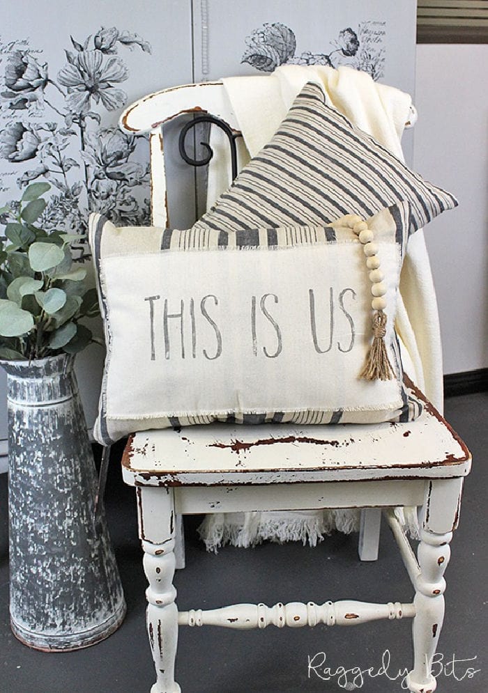 A DIY Farmhouse stamped tea towel that says THIS IS US, made into a oblong shaped cushion with grey striped fabric. The cushion is sitting on a white chippy painted chair, with a second cushion and a cream coloured throw behnd it.