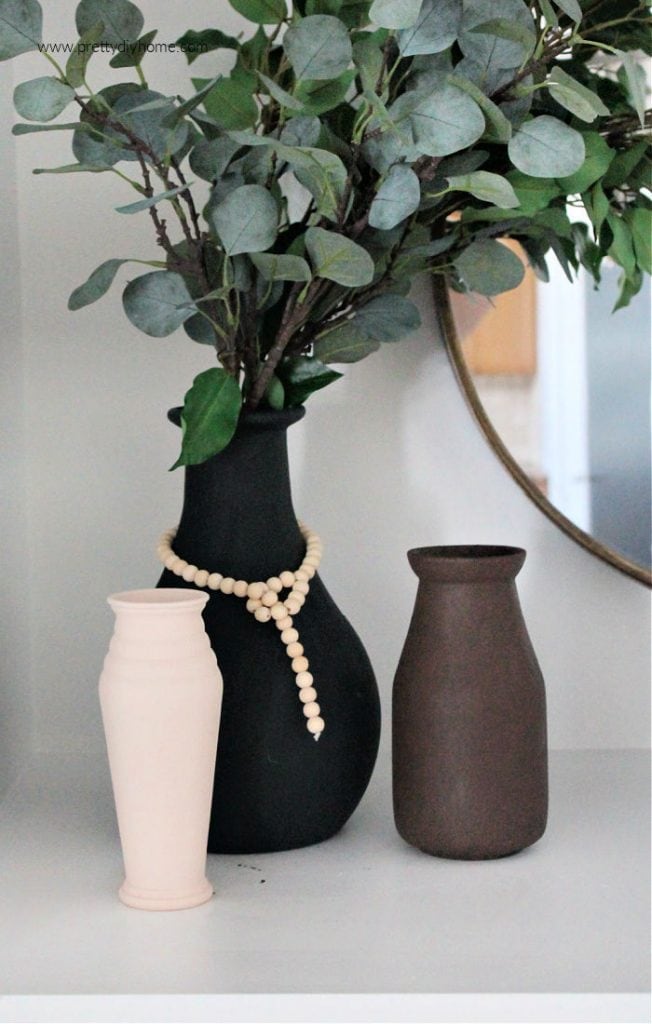 Three different DIY faux ceramic vases on a mantel. One vase is black, one is brown, and one is cream coloured. The black vase is the largest with a wooden farmhouse string of beads and has branches of greenery. They are all sitting beside a round mirror with metal frame.