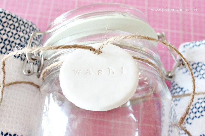 A glass jar being held in place by two towels and have a polymer clay DIY tag attached to the front with twine.