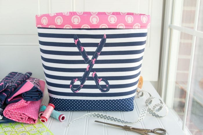 A large fabric bin with a appliqued pair of scissor on the front. The basket has a blue base, striped fabric and handmade pink brim. An easy to make large fabric basket tutorial to sew.