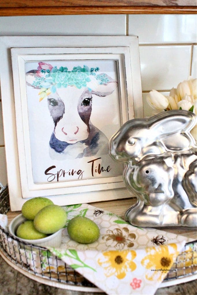 A farmhouse cow Spring printable in a kitchen arrangement. The printable has a black and white calf with a succulent floral arrangement on its head. The heading of the printable says Spring time. The printable is part of a Spring kitchen arrangement with white tulips, an antique rabbit chocolate form, and soft green Easter eggs.