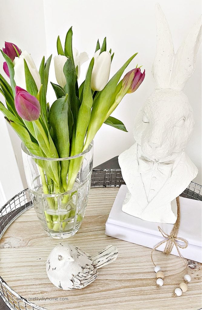 A fresh bouquet of tulips in bright colours that have been prepared so that they last longer. The fresh tulips are in a nice real crystal vase and their is white Easter farmhouse decor surrounding it. A little white bird, white wrapped books and a white sculptured bunny.