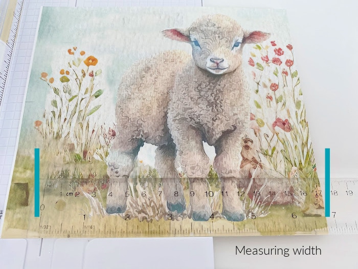 A spring artwork of a baby lamb standing among flowers is being cut to a specific size. The image shows a ruler with markings for the measurement.