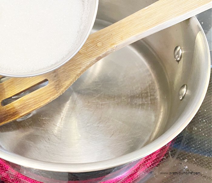 Making simple syrup in a small saucepan. There is a pan of water on the head with a wooden spoon and sugar being poured into it.