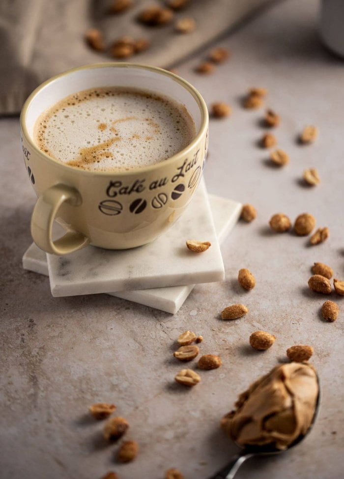 A small mug filled with a creamy peanut butter latte coffee recipe.  The mug is small and has foam on top.  The background has peanuts and a spoon of peanut butter.