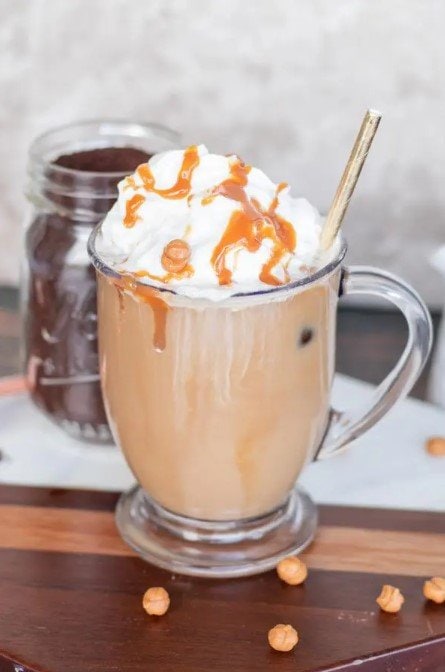 An iced caramel macchiato coffee served in a clear glass mug with whipped cream topping and caramel sauce. The background has a jar of dark fresh coffee grounds and there are caramel chips scattered on the counter.