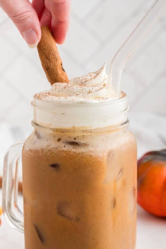 A large icy mug filled with iced pumpkin latte coffee.  The coffee is topped with whipped cream and is being stirred with a cinnamon stick.