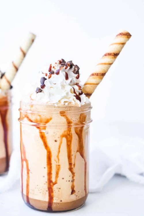 A mason jar filled overflowing with a mocha Frappuccino. The mocha Frappuccino is topped with whipping cream and chocolate sauce. and garnished with a Pirouline cookie stick.