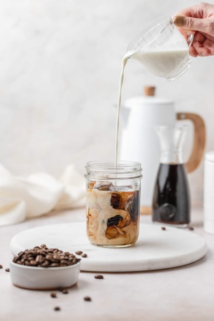 A mason jar glass filled with cold brew coffee thats having cream poured into it.