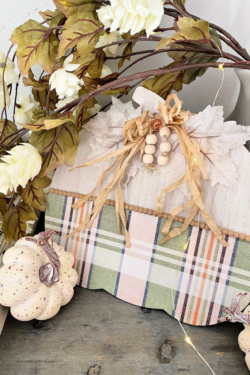 A dollar tree pumpkin decorated with a pastel orange and green plaid, white farmhouse beads, and cream coloured half beads. The pumpkin is part of a fall decor vignette.
