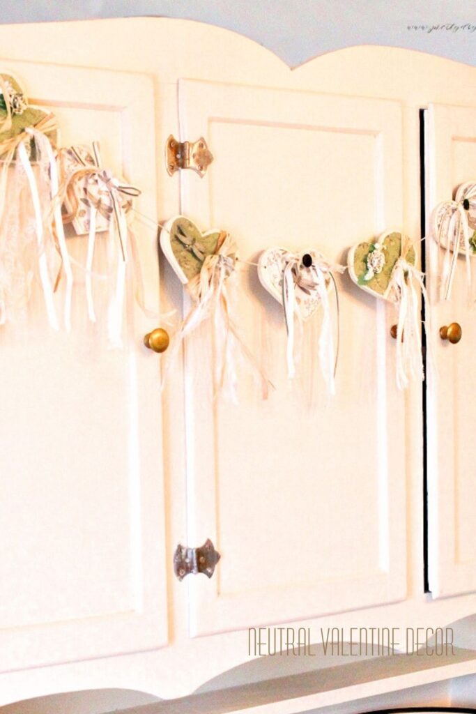 DIY wood Valentine’s Day decor featuring a sage green and white upcycled wood heart banner hanging on a cream antique cabinet.