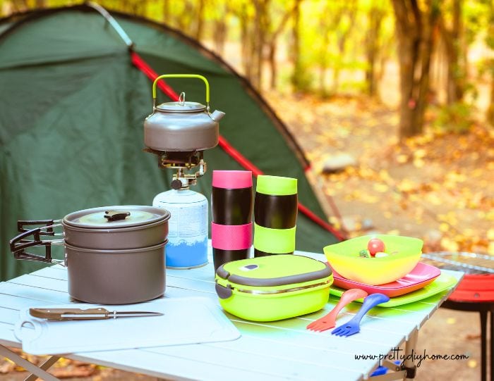 A campsite with a table covered in cheerful colored camp cooking equipments a pot, insulated mugs, plastic cotainer.