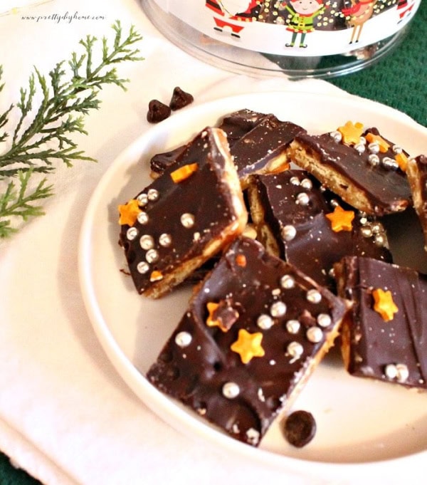 A white plate covered with chocolate cover Christmas crack toffee. The chocolate toffee has silver and gold Christmas sprinkesl on top and i, and the background is decorated with a Christmas tablecloth and greenery.