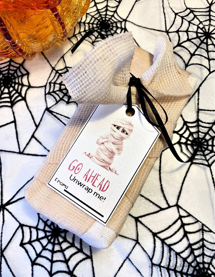 A box wrapped up in bandage on a Halloween spiderweb background. the bandage wrapped gift box has a mummy tag on top that says Go Ahead Unwrap Me.