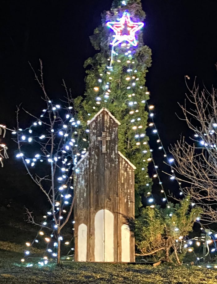 An outdoor Christmas decor display featuring a rustic wood church decoration, and lots of sparkling white and cream lights.