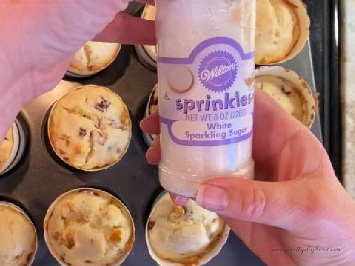 An image of coarse white sugar in a plastic bottle.  The bottle is held over a pan of freshly out of the oven panettone muffins.