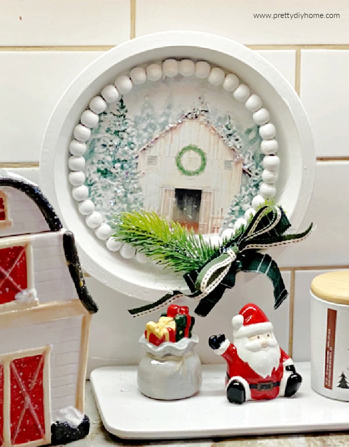 A white painted  Christmas kitchen sign made using a dollar tree pie plate painted white and wood beads and a lovely free printable barn print of a white barn at Christmas.