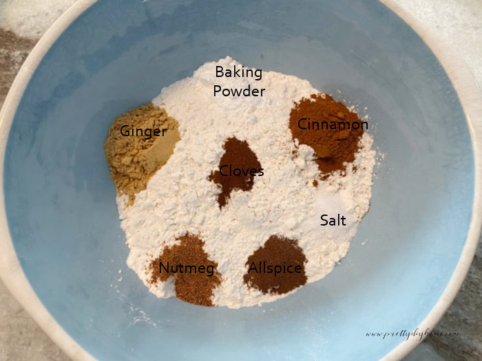 A large mixing bowl filled with dry ingredients and assorted spices for making gingerbread scones. Each ingredient is labeled with text, ginger, nutmeg, cinnamon etc.