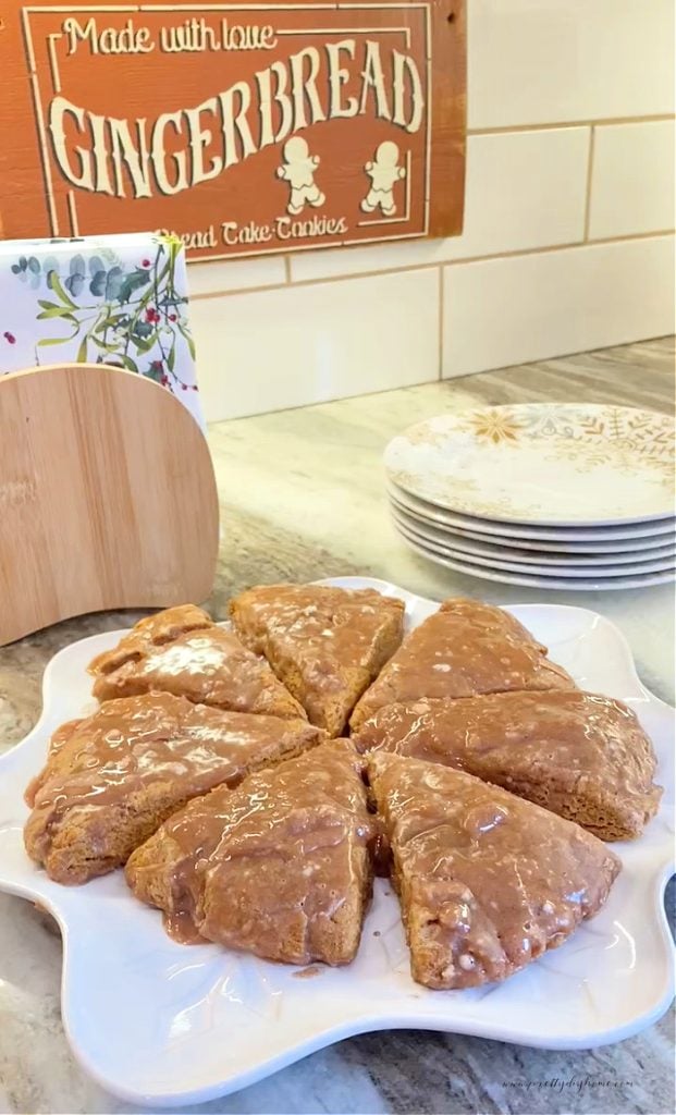 A white snowflake shaped Christmas tray covered with a circle of 8 gingerbread sccones, each with shiny cinnamon glaze.  There is a stack of Christmas plates and napkins in the background.