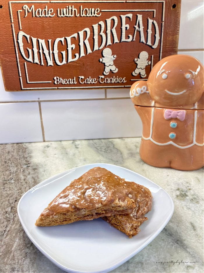 Homemade gingerbread scones with a glossy cinnamon glaze. The ir are two scone wedges on a white plate with a gingerbread man cookie jar in the background.