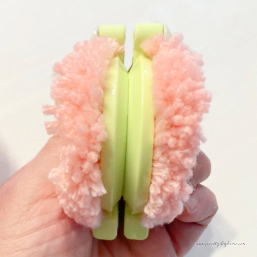 A side view of a green pom pom maker you can see the groove in the middle and the cut pink fluffy yarn strings of the pom pom sticking out both sides.