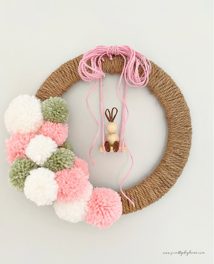 An Easter wreath DIY made with large fluffy pink white and green Pom Poms, and a handmade beaded Easter bunny hanging from a swing. The wreath is covered in brown twine and the bunny is hanging from a DIY swing made using pink twine.